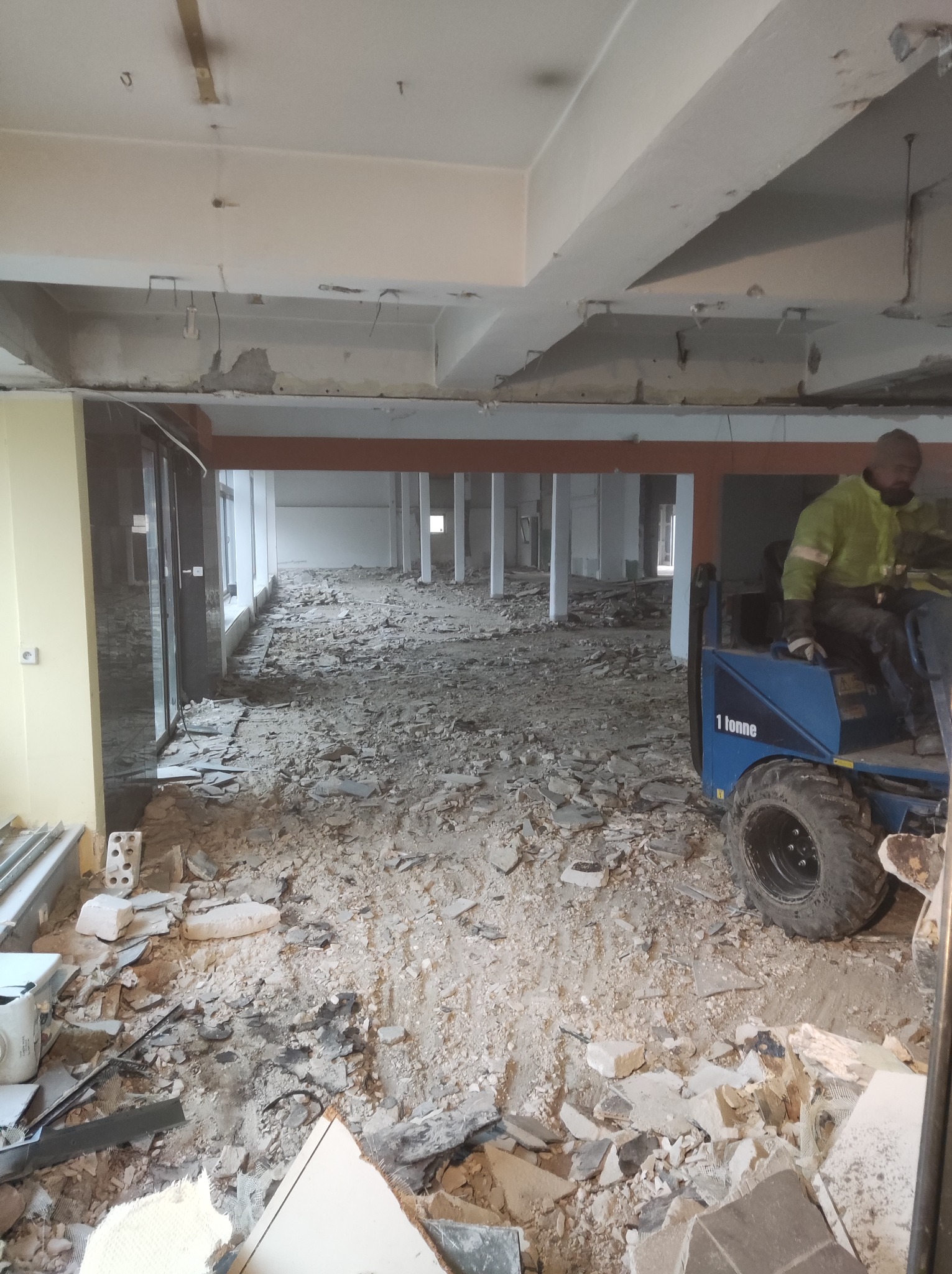 Wnętrze budynku w trakcie rozbiórki, gruz na podłodze, widoczny pracownik na niebieskim minitraktorze 1 tonne, odsłonięte elementy konstrukcyjne sufitu.