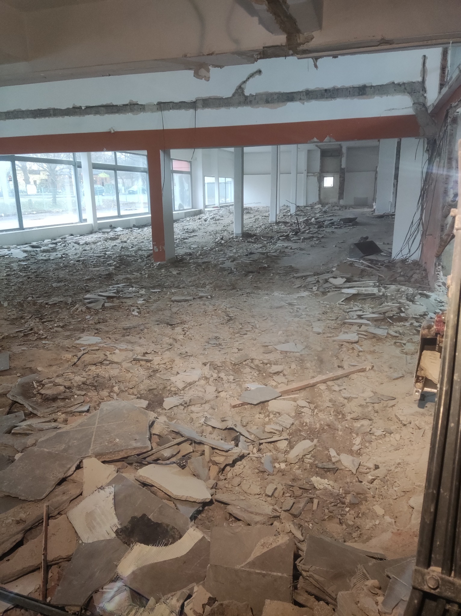 Wnętrze budynku w trakcie rozbiórki, widoczne gruzowisko na podłodze, odsłonięte elementy konstrukcyjne sufitu i ścian, duże okna w tle.