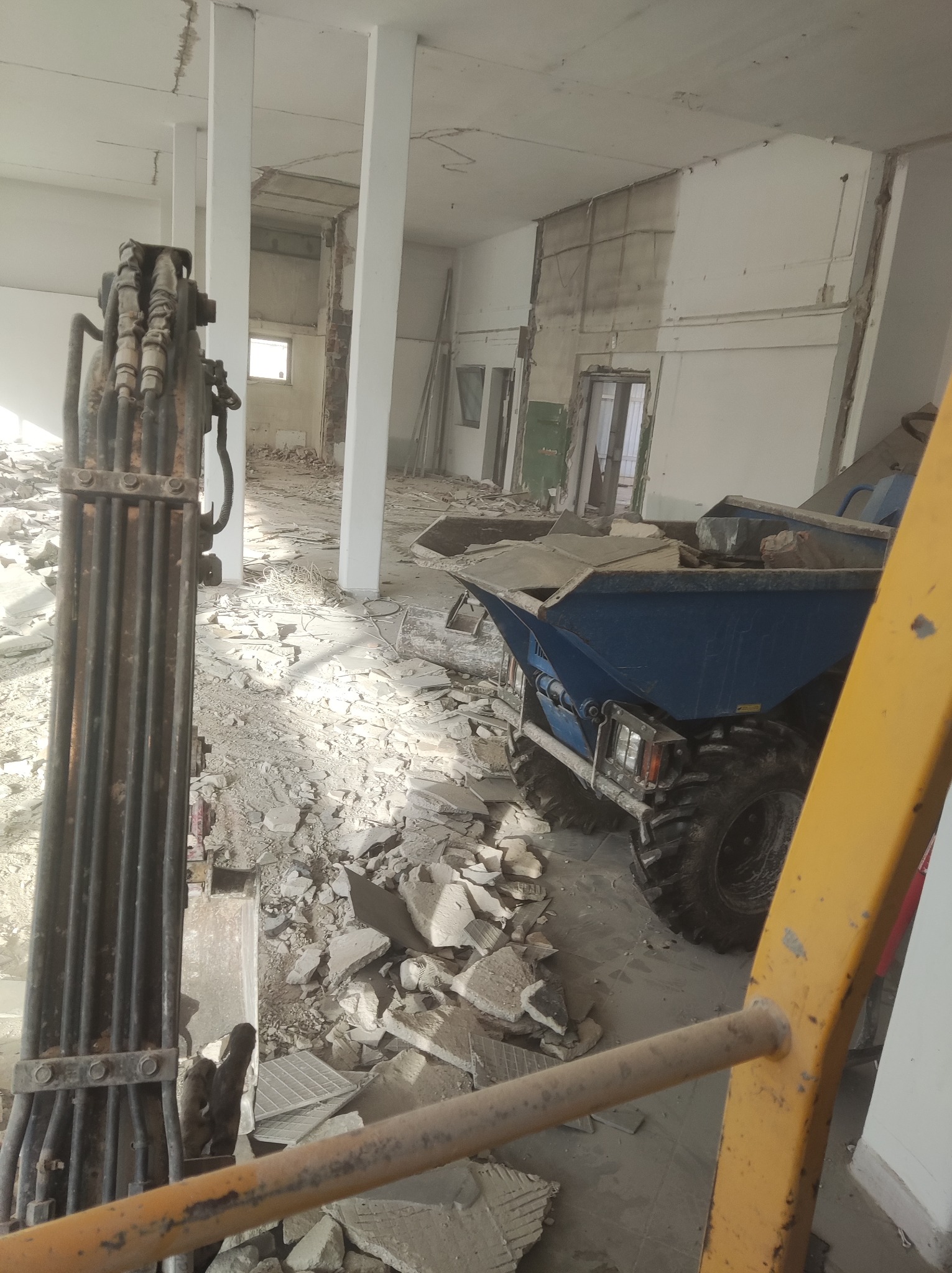 Wnętrze budynku w trakcie rozbiórki, widoczne fragmenty gruzu, maszyna budowlana i elementy konstrukcyjne, oświetlone naturalnym światłem wpadającym przez okno.