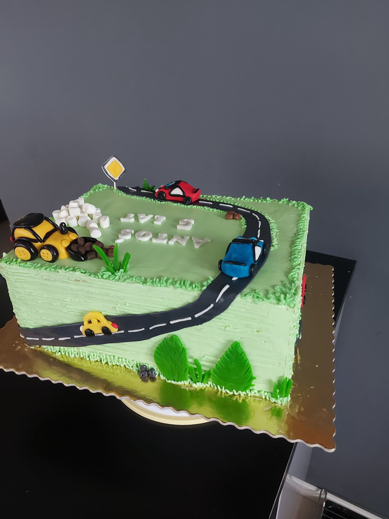 Kwadratowy tort urodzinowy z jasną, zieloną polewą imitującą trawę, dekorowany czarną drogą z białymi pasami, modelami samochodów (żółty, czerwony, niebieski), znakiem drogowym, marshmallow...