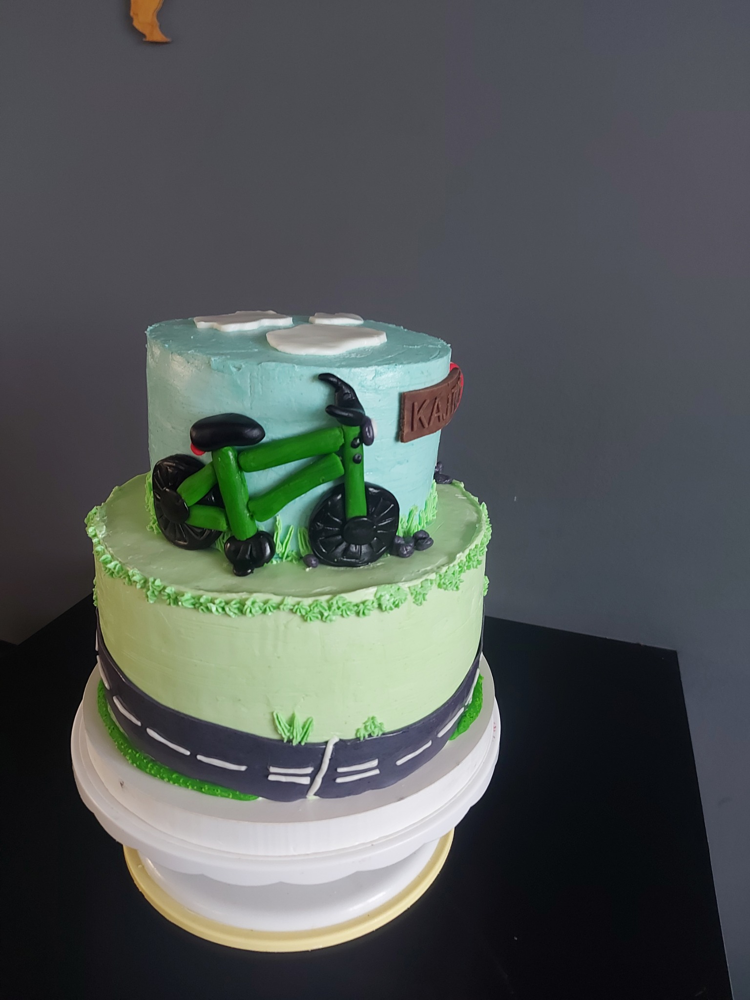 Dwupoziomowy tort w Lublinie z motywem rowerowym: zielony rower z lukru na tle błękitnego nieba z obłokami i drogą dookoła podstawy.