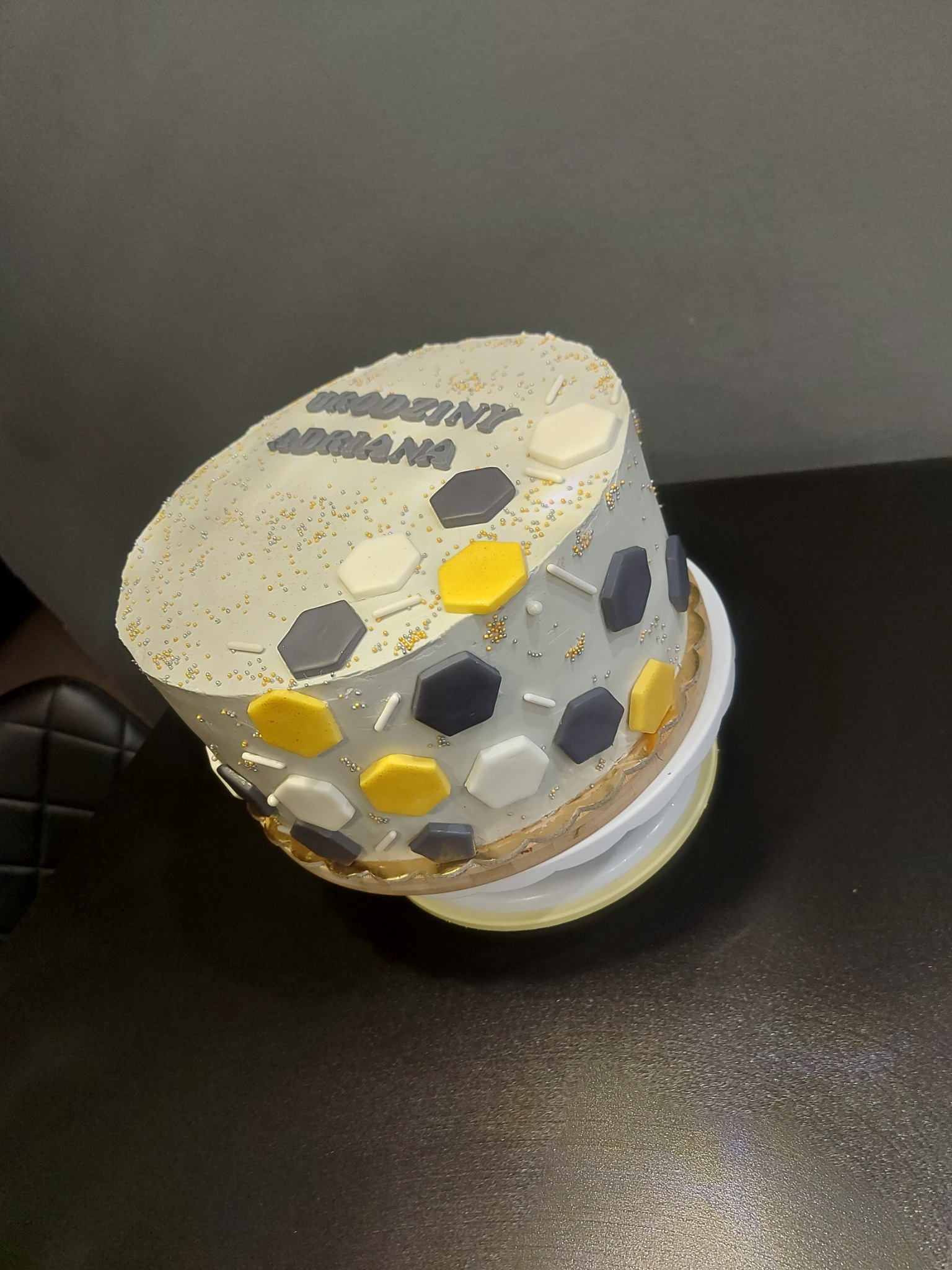 Tort urodzinowy dla Adriany z dekoracjami w kształcie heksagonów w kolorach białym, szarym i żółtym, posypany drobnymi, złotymi kuleczkami.