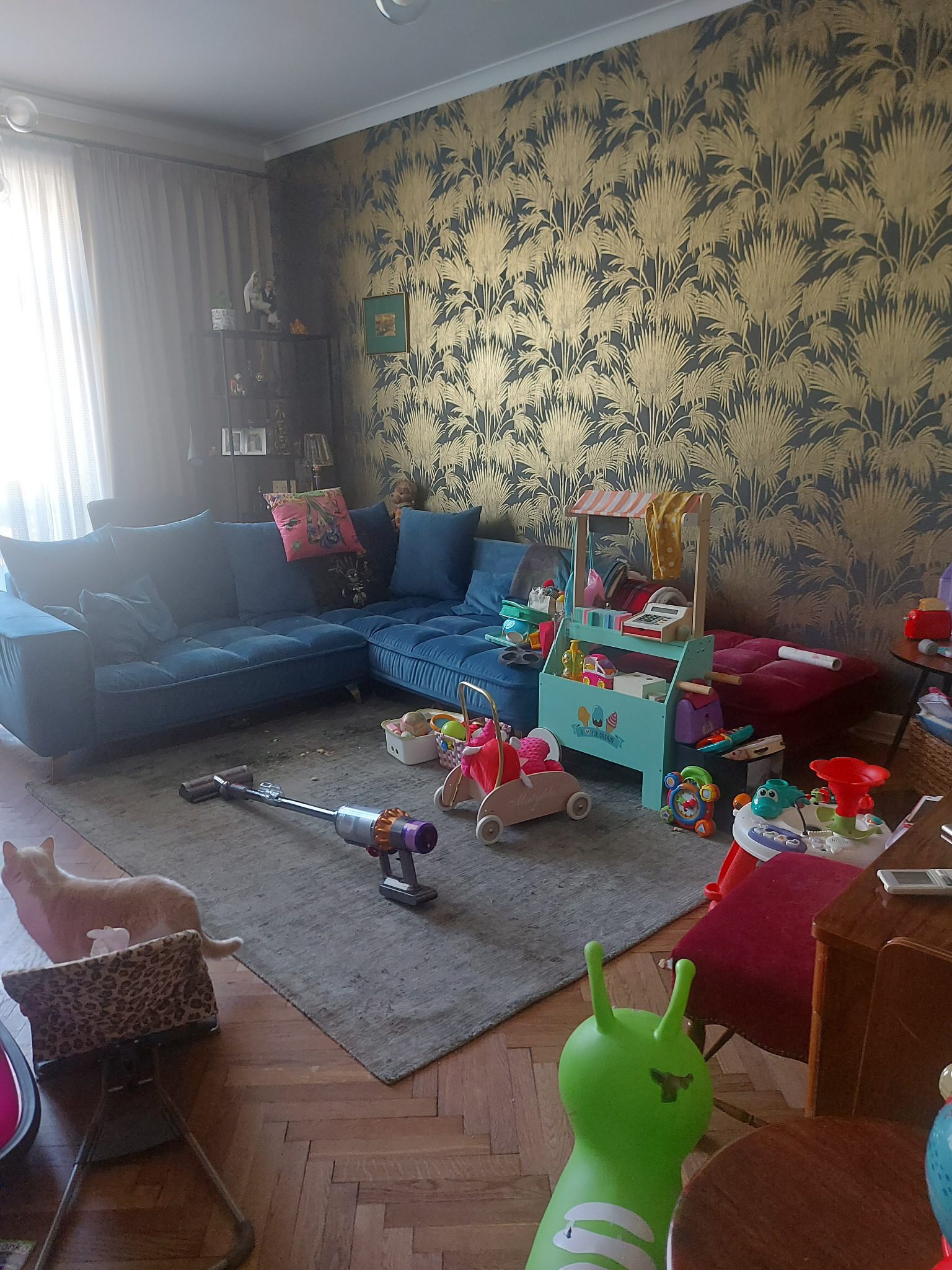 Wnętrze pokoju dziennego z niebieską sofą narożną, wzorzystą tapetą i rozrzuconymi zabawkami dziecięcymi; odkurzacz pionowy na dywanie w trakcie użycia.