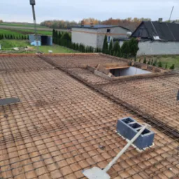 Zbrojenie stropu domu w trakcie budowy. Widoczna siatka z prętów stalowych, drewniane szalunki i narzędzia. W tle zabudowa jednorodzinna i krajobraz.