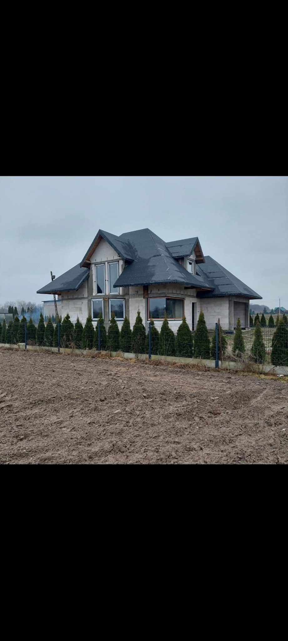 Surowy dom jednorodzinny z ciemnym dachem i dużymi oknami, ogrodzony niskim płotem i żywopłotem. Widok na plac budowy w pochmurny dzień.