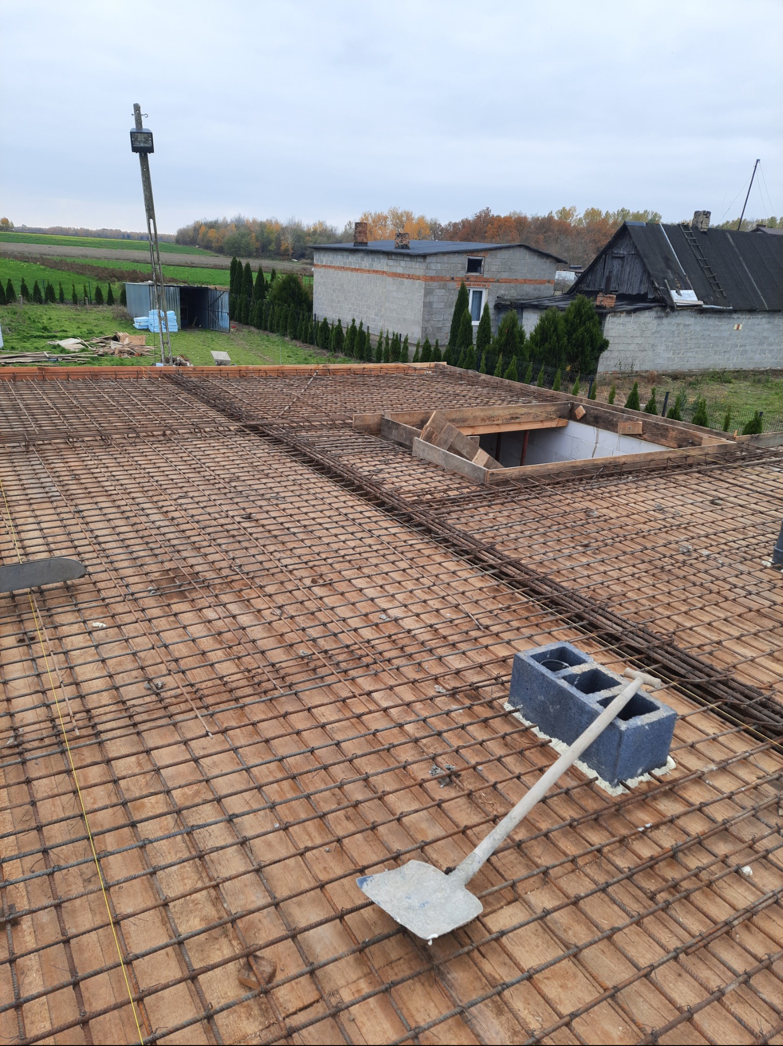 Zbrojenie stropu domu w trakcie budowy. Widoczna siatka z prętów stalowych, drewniane szalunki i narzędzia. W tle zabudowa jednorodzinna i krajobraz.