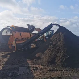 Pomarańczowa przesiewarka mobilna Donnstadt SM-518 przesypuje ziemię na dużą pryzmę, widoczny operator w kabinie, błękitne niebo w tle.