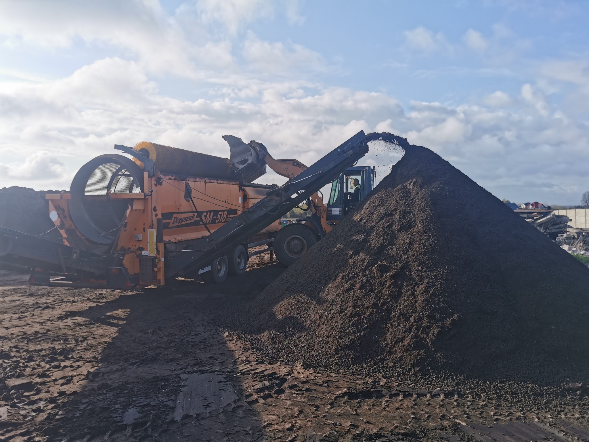 Pomarańczowa przesiewarka mobilna Donnstadt SM-518 przesypuje ziemię na dużą pryzmę, widoczny operator w kabinie, błękitne niebo w tle.