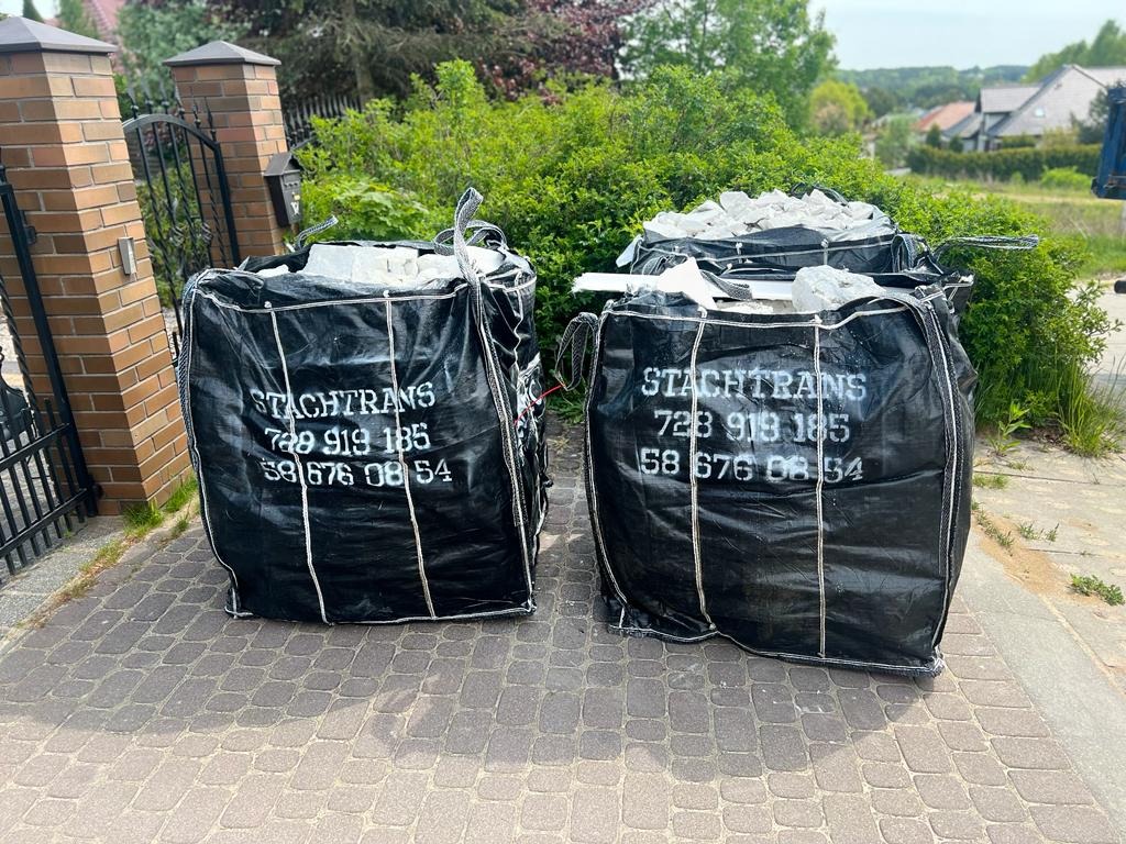 Dwa czarne, worki typu big bag z białym nadrukiem 'STACHTRANS' wypełnione gruzem, stojące na kostce brukowej przed posesją z zielenią w tle.