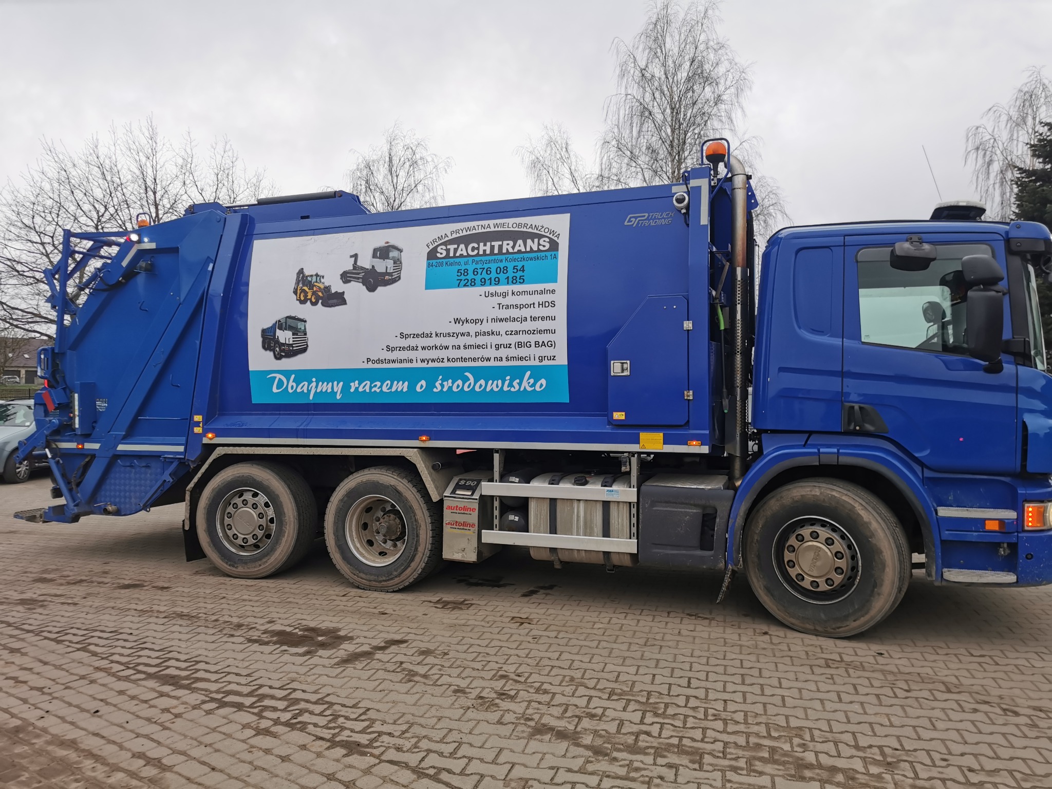 Niebieska śmieciarka z reklamą firmy STACHTRANS, oferującej usługi komunalne, transport HDS oraz sprzedaż i wywóz kontenerów na śmieci i gruz, zaparkowana na kostce brukowej.