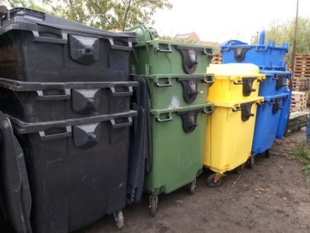 Rząd kolorowych kontenerów na kółkach w Gdyni: czarne, zielone, żółte i niebieskie. Kontenery ustawione na nierównym, ziemistym podłożu, z widocznymi paletami w tle.