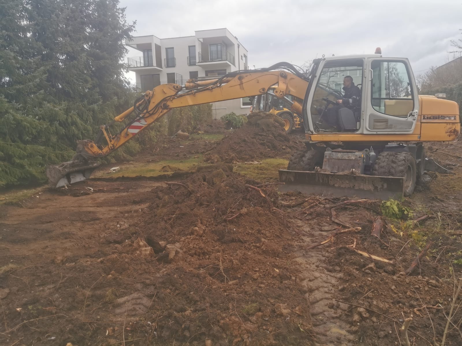 Żółta koparka Liebherr na placu budowy, w tle budynek mieszkalny i druga maszyna, widoczne świeżo przekopane błotniste podłoże.