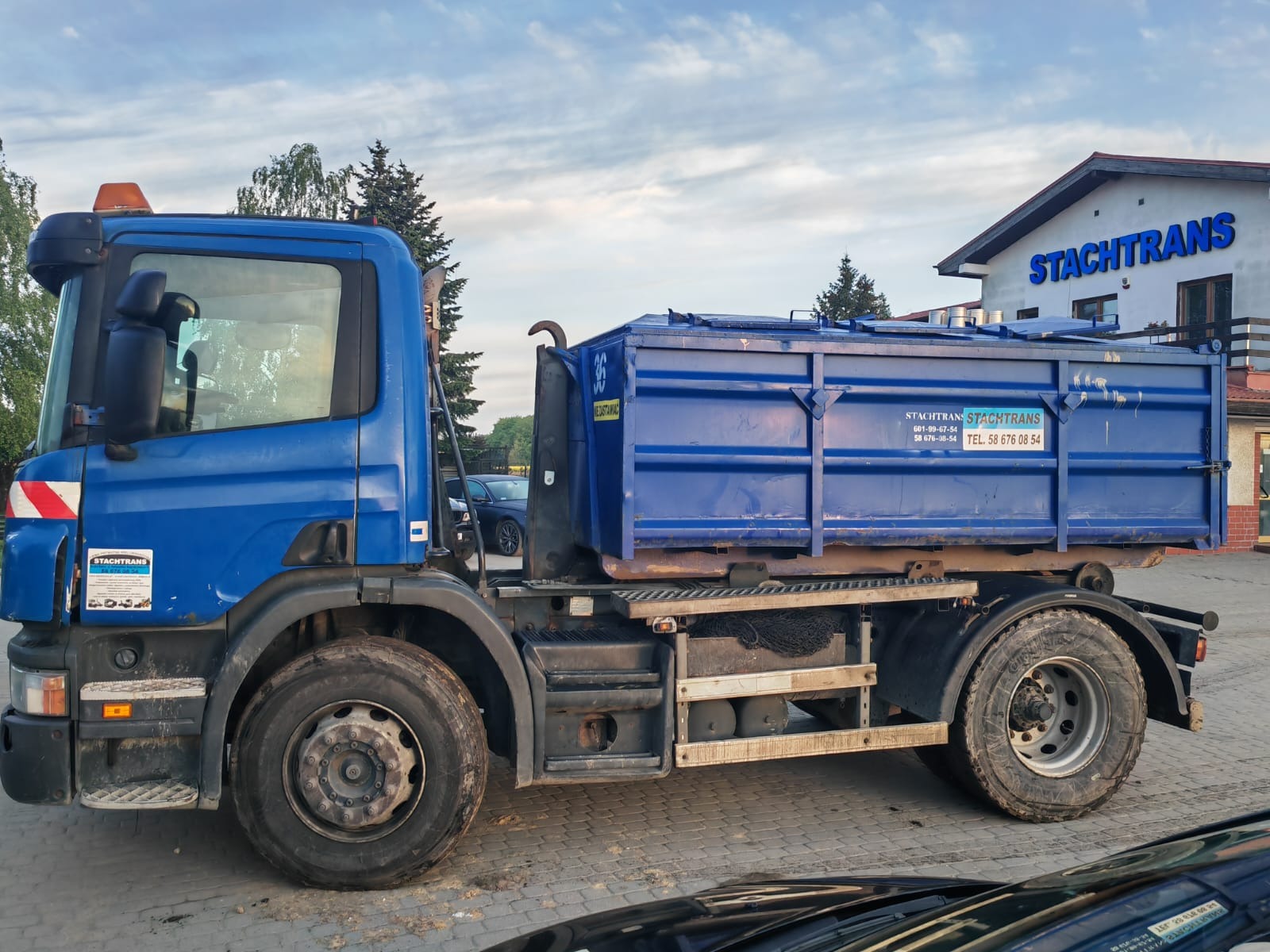 Niebieska ciężarówka z kontenerem, z widocznym logo firmy transportowej Stachtrans i numerem telefonu, zaparkowana na brukowanym placu przed budynkiem firmy.