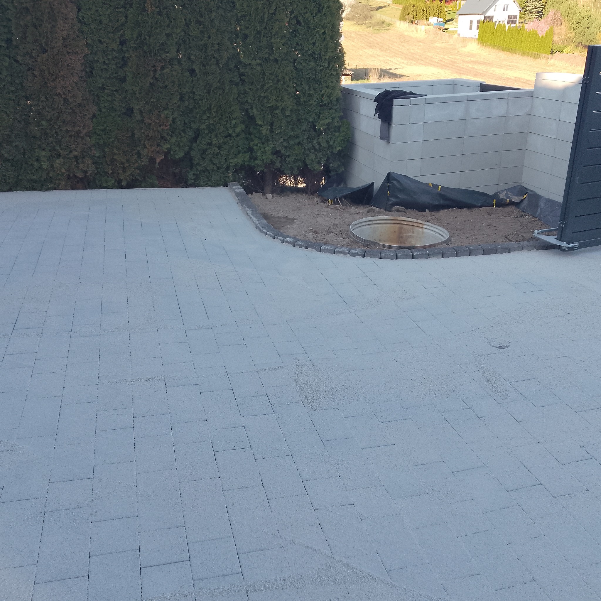 Jasnoszara kostka brukowa ułożona na posesji, obok zielony żywopłot i betonowe elementy. Widoczny właz kanalizacyjny, teren częściowo przygotowany pod nasadzenia.