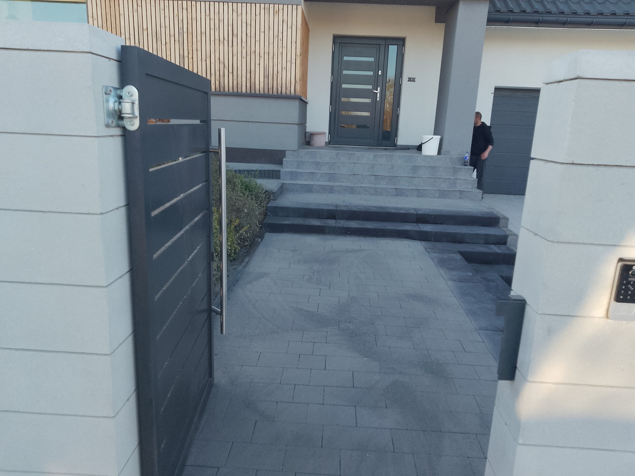 Nowoczesne wejście do domu w Rzeszowie z szarej kostki brukowej i metalową bramą. Schody prowadzą do drzwi wejściowych, widoczny fragment elewacji.