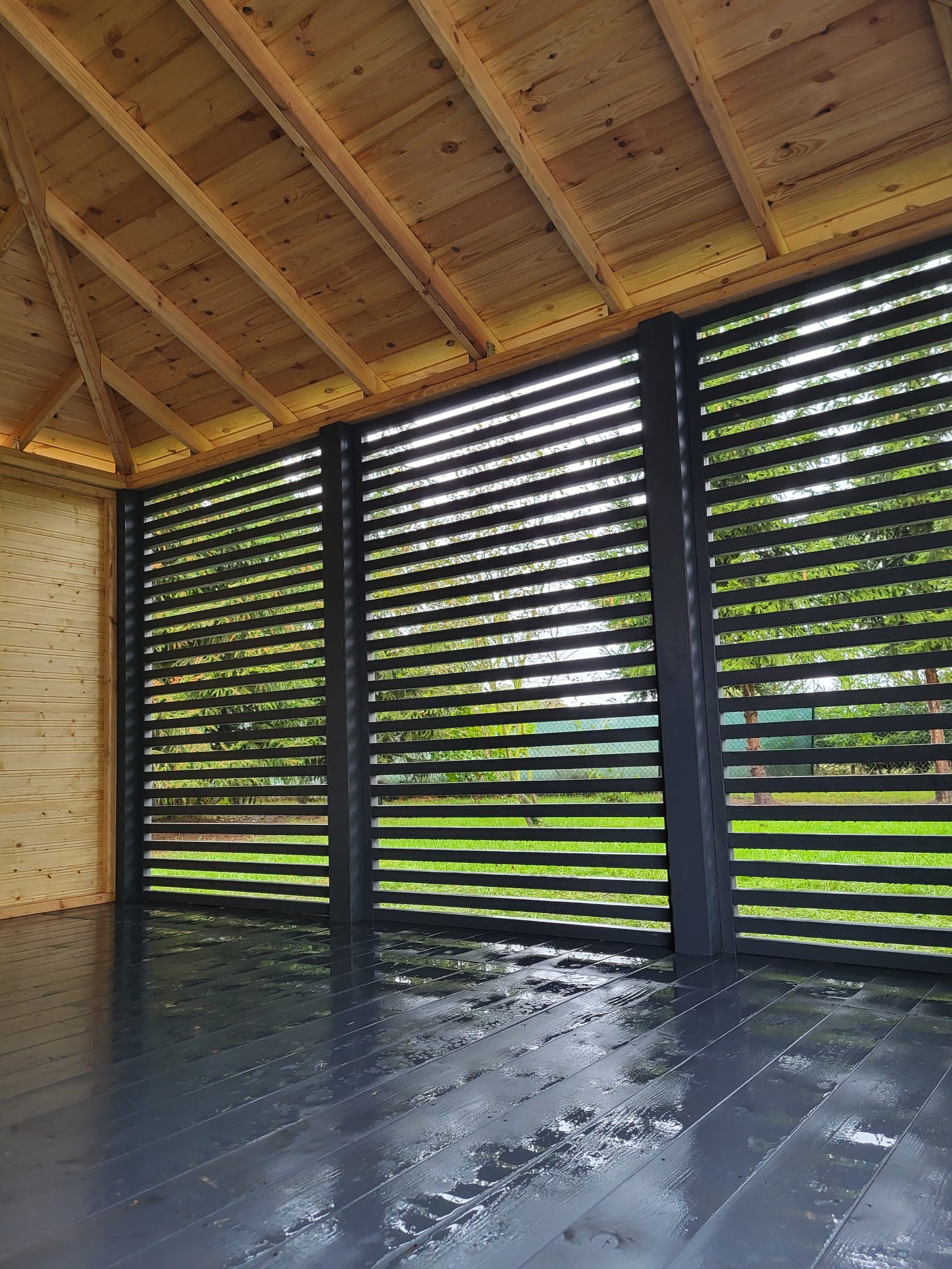 Wnętrze drewnianej konstrukcji z ażurowymi, poziomymi przesłonami w kolorze grafitowym, widok na zewnątrz z zielenią i drzewami, mokra, błyszcząca podłoga z desek w kolorze grafitowym, drewniany...