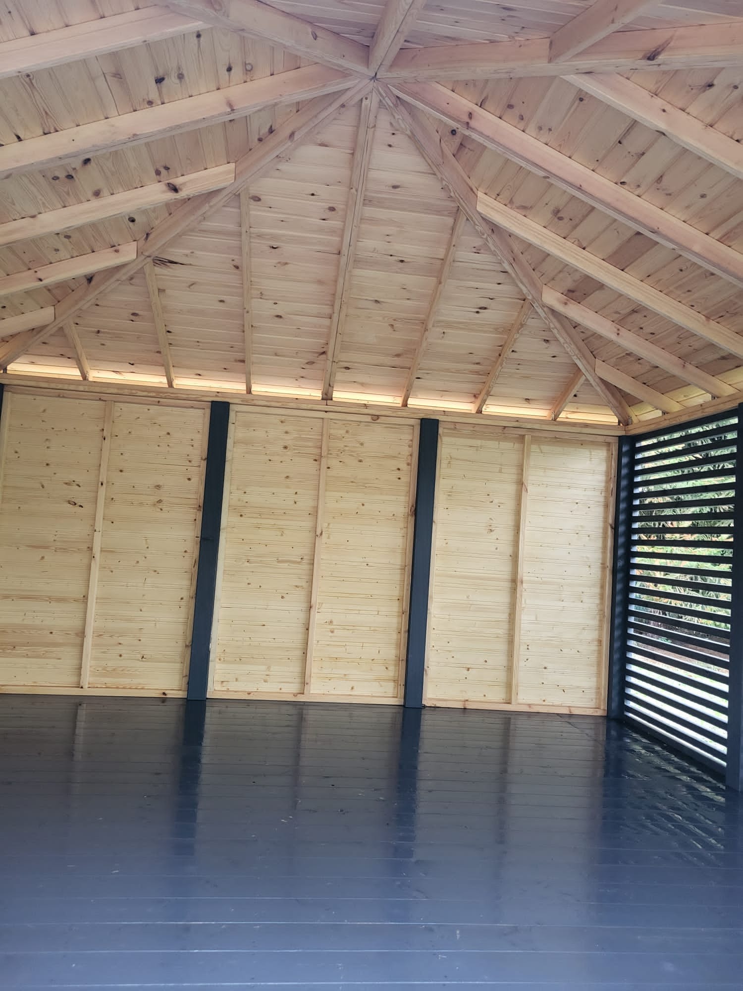 Wnętrze altany z drewnianą konstrukcją dachu, ścianami z jasnego drewna i ciemną podłogą, widok z dołu.