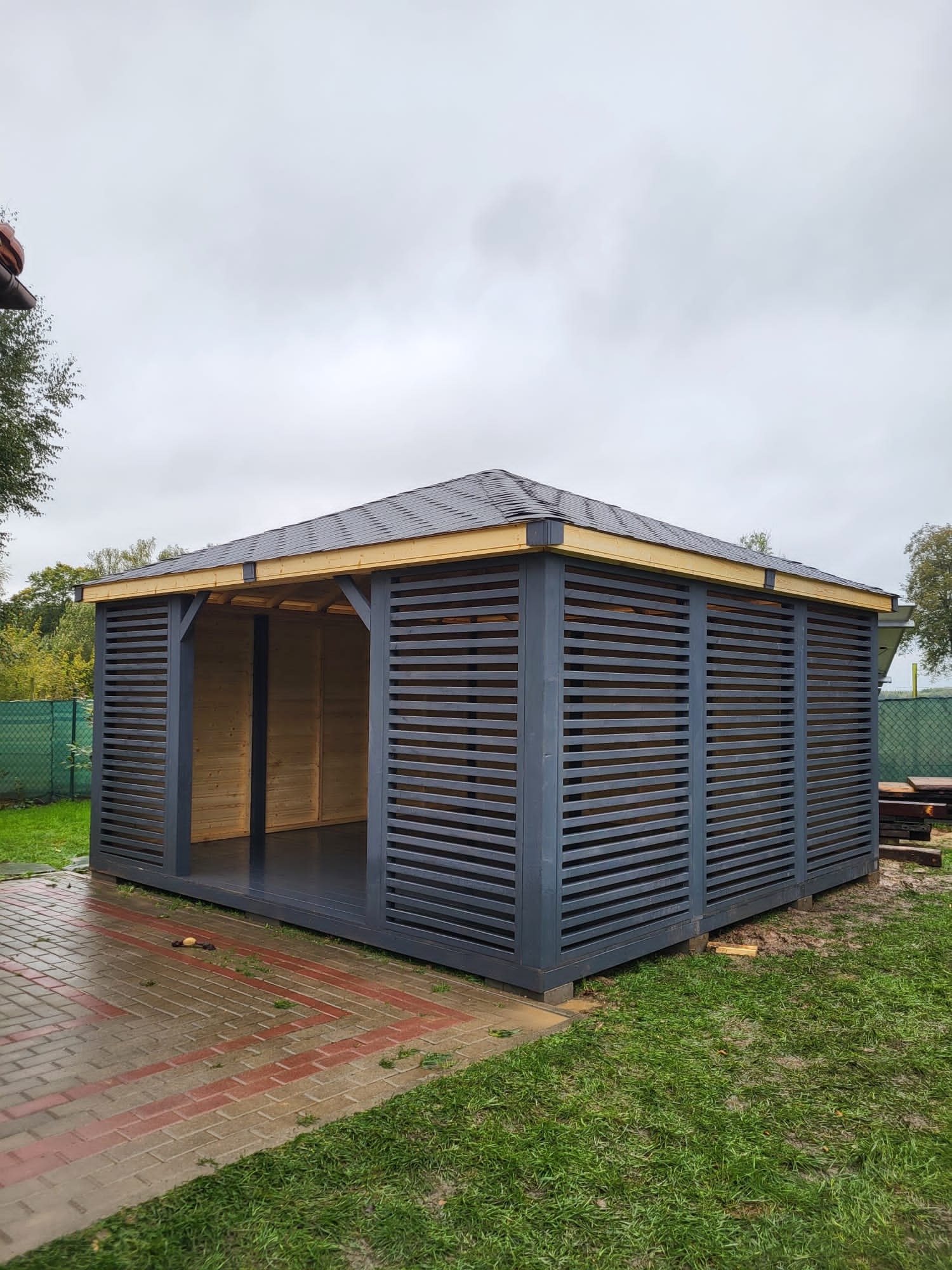 Szara, drewniana konstrukcja z poziomymi listwami i dachem z papy bitumicznej, otoczona trawnikiem i brukowanym chodnikiem, w pochmurny dzień.