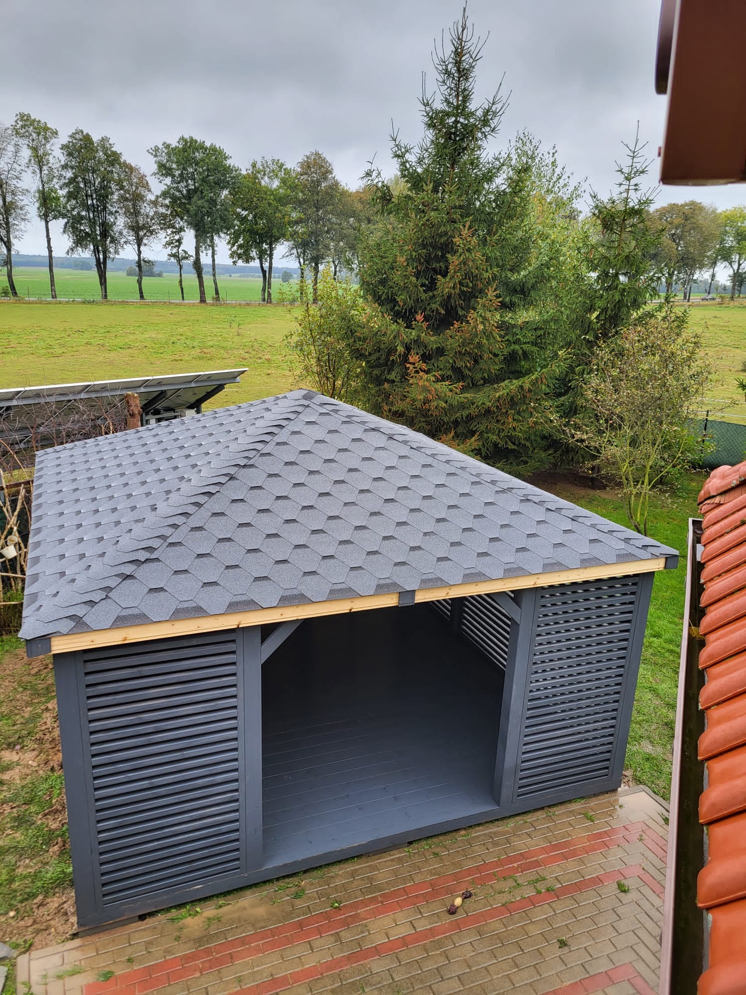 Nowoczesna altana z szarą dachówką bitumiczną i lamelowymi ścianami, widok z góry na tle zielonego pola i drzew
