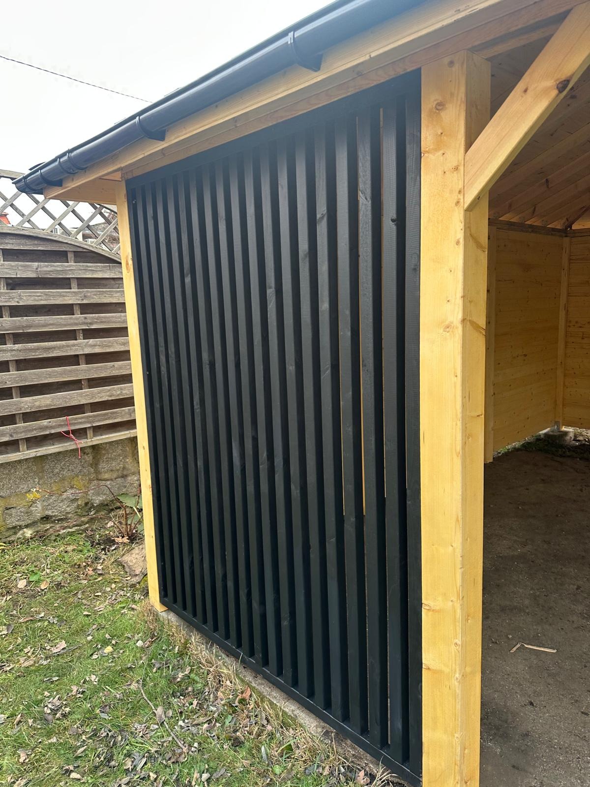 Fragment altany 5x4 z czarnymi, pionowymi deskami tworzącymi ścianę, widoczny z zewnątrz, z fragmentem dachu i słupem konstrukcyjnym z surowego drewna.