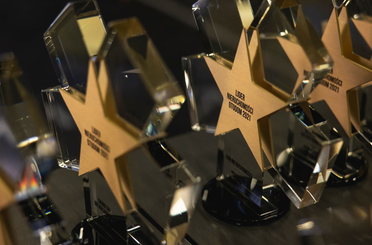 Szereg nagród w kształcie gwiazdy z napisem 'Lider Nieruchomości Otodom 2021' na czarnych podstawkach, wykonanych z przezroczystego szkła i złotego metalu.
