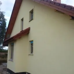 Jasnożółta elewacja domu z brązowym dachem. Widoczne okna z brązowymi ramami i fragment ogrodu z drzewami. Ujęcie z boku budynku, w tle błękitne niebo.