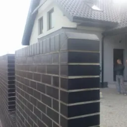 Nowoczesny, ciemnobrązowy mur z cegły klinkierowej, tworzący ogrodzenie posesji. W tle dom z szarą elewacją i dwuspadowym dachem. Dwóch mężczyzn stoi obok wejścia.