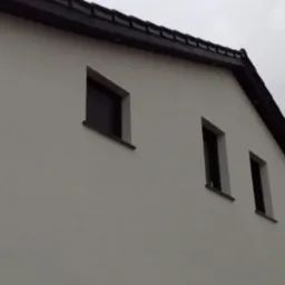 Fragment elewacji domu z czterema oknami, jedno z opuszczoną roletą, pozostałe bez, pod ciemnym dachem z dachówki ceramicznej, pochmurne niebo w tle.