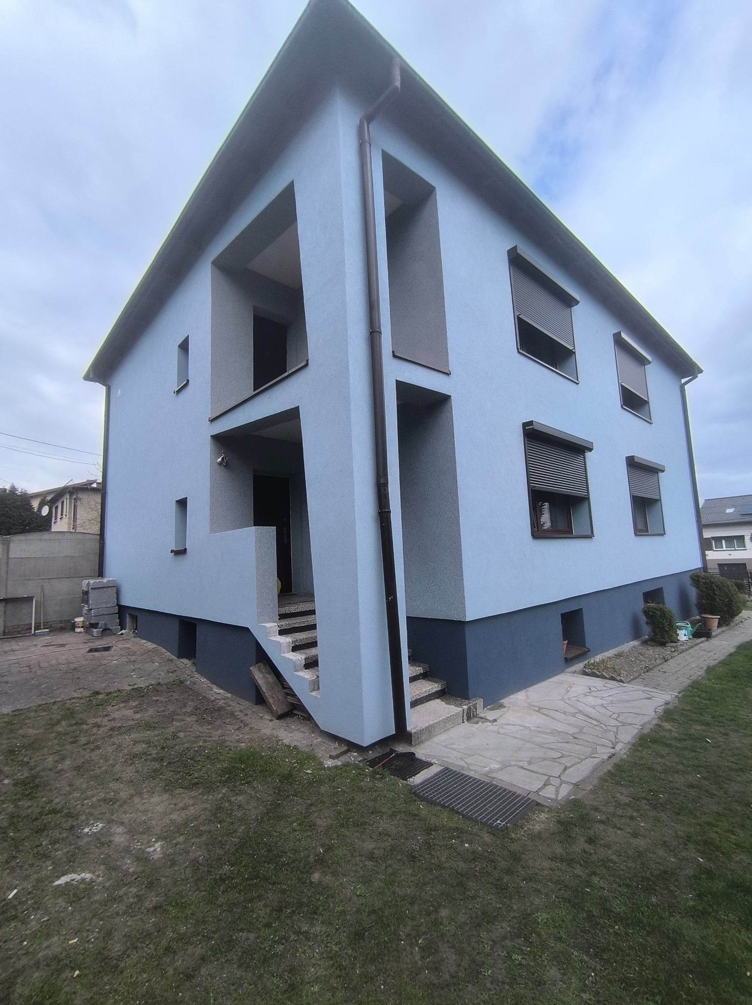 Niebieski dom po remoncie elewacji z ciemnoszarym cokołem i roletami w oknach. Widoczne schody wejściowe i elementy wykończenia.
