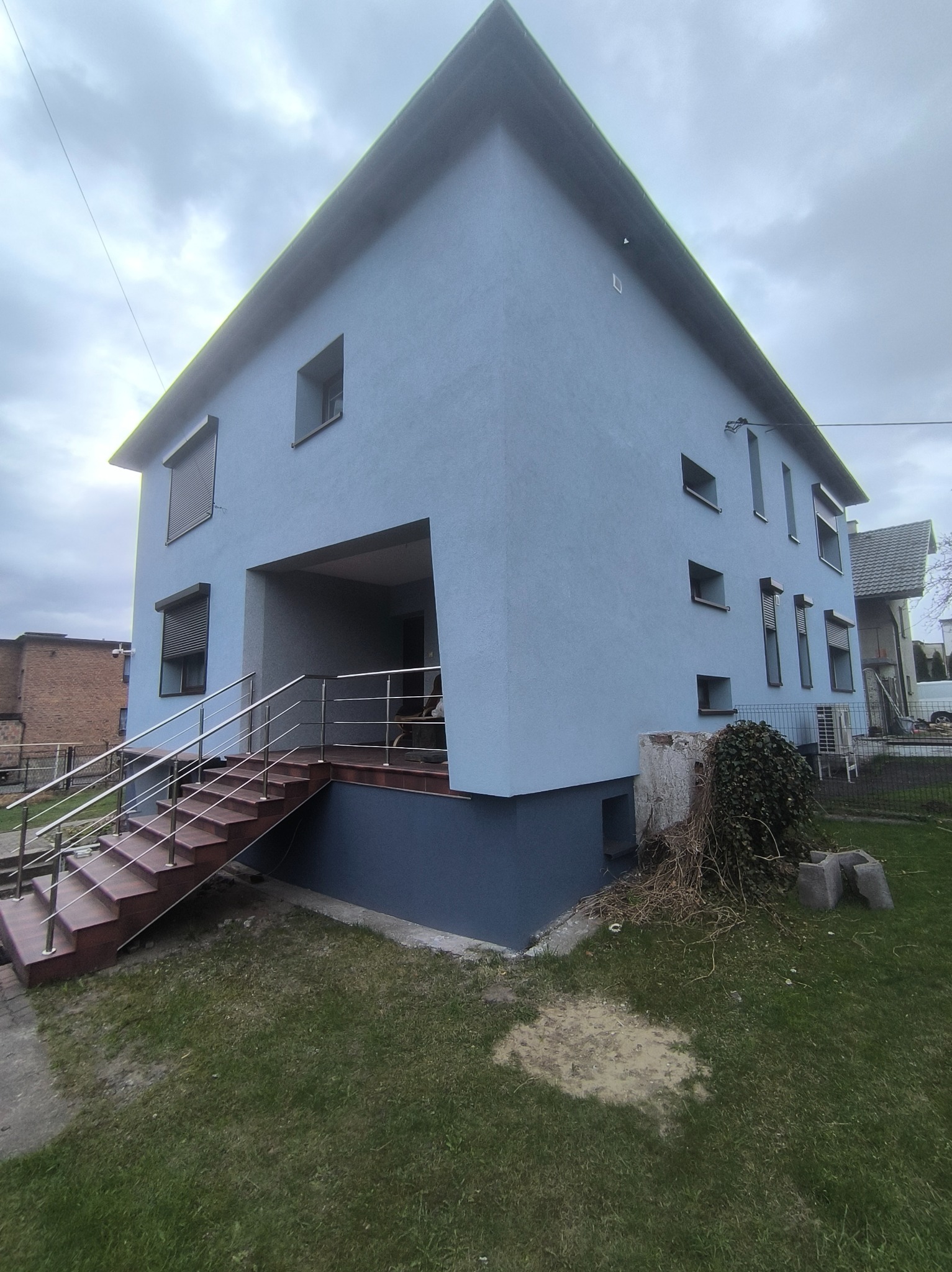 Narożne ujęcie dwukondygnacyjnego domu z niebieską elewacją i metalową balustradą schodów wejściowych, na tle pochmurnego nieba.