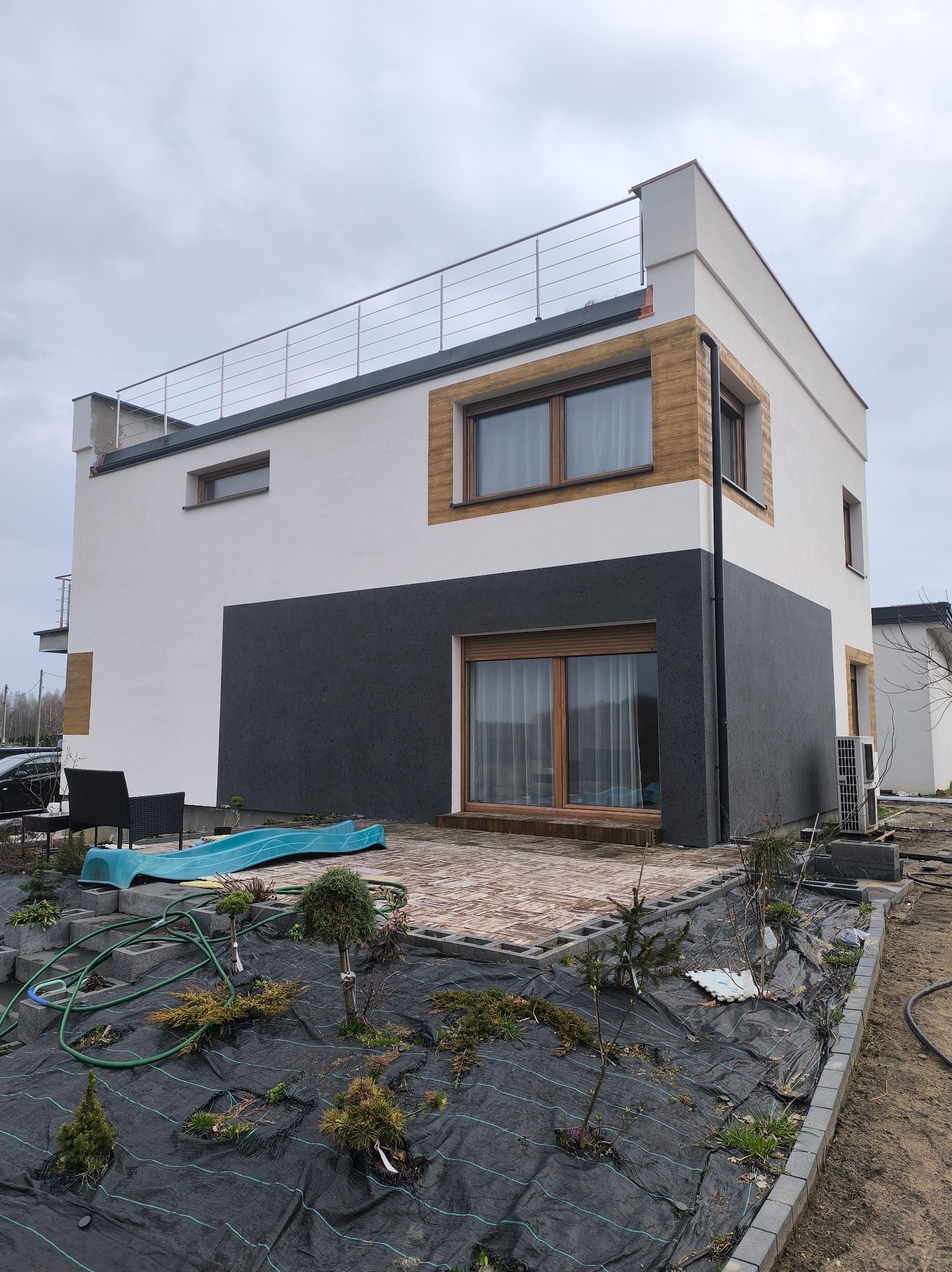 Dwukondygnacyjny dom z płaskim dachem i drewnianą elewacją, z szarą dolną częścią i białym górnym piętrem, widziany z perspektywy ogrodu w pochmurny dzień.