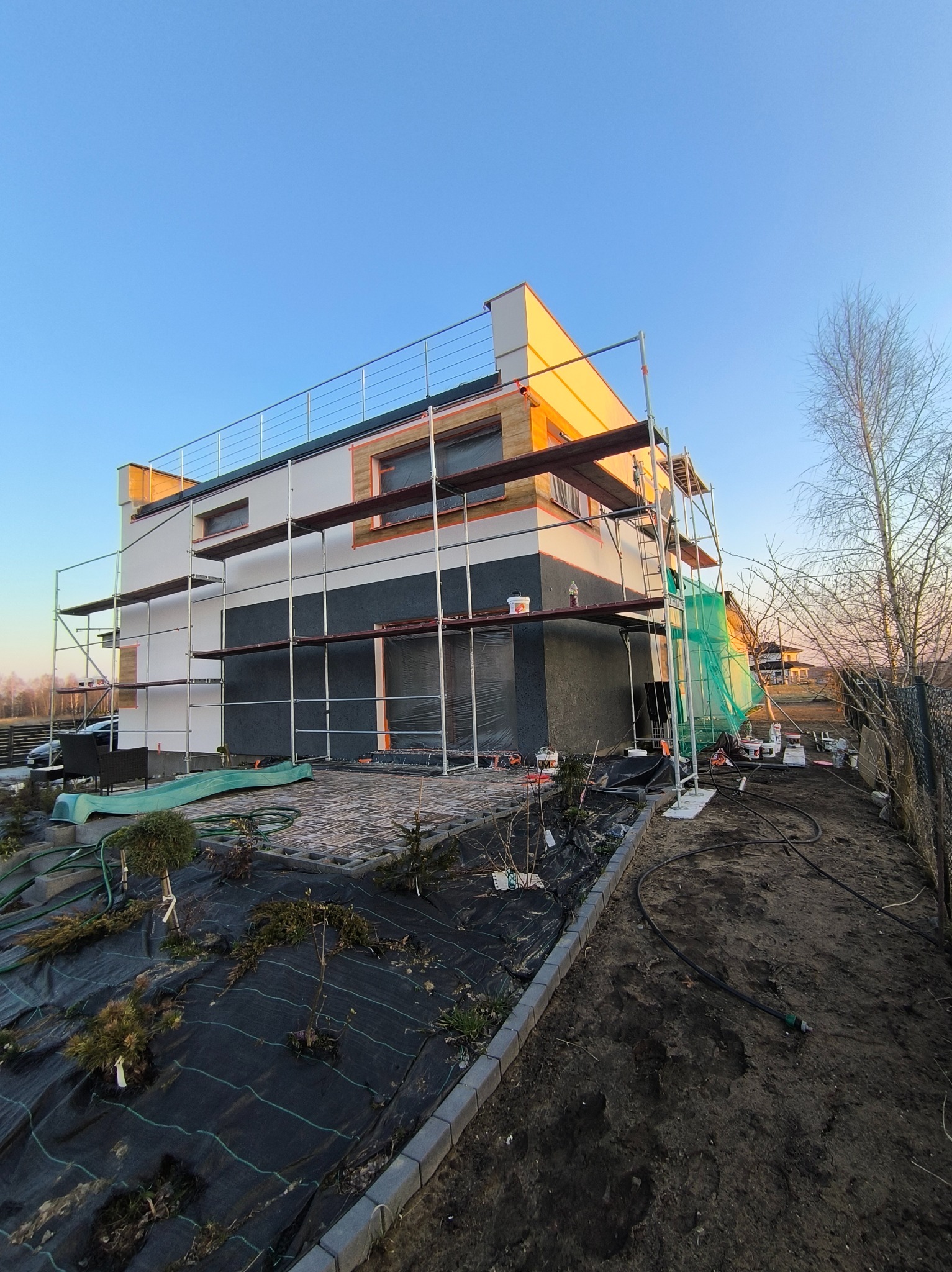 Dom w trakcie prac elewacyjnych, otoczony rusztowaniami. Widoczne fragmenty ocieplenia, puszki z farbą i narzędzia. Prace w toku, zieleń w ogrodzie w tle.