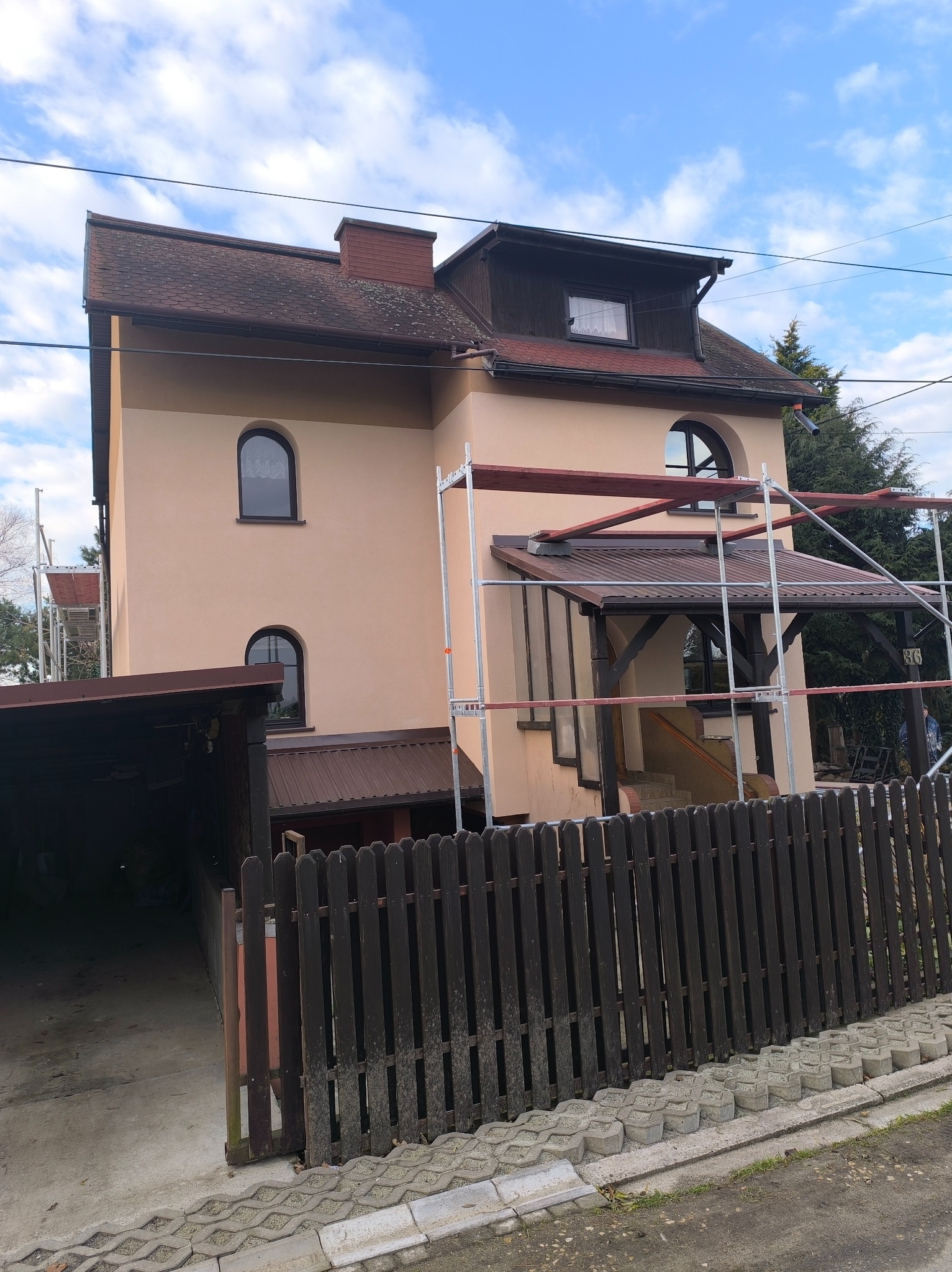 Dom z beżową elewacją w trakcie prac. Widoczne rusztowania, brązowy dach i okna w kształcie łuków. Uwagę zwraca drewniany płot i błękitne niebo w tle.