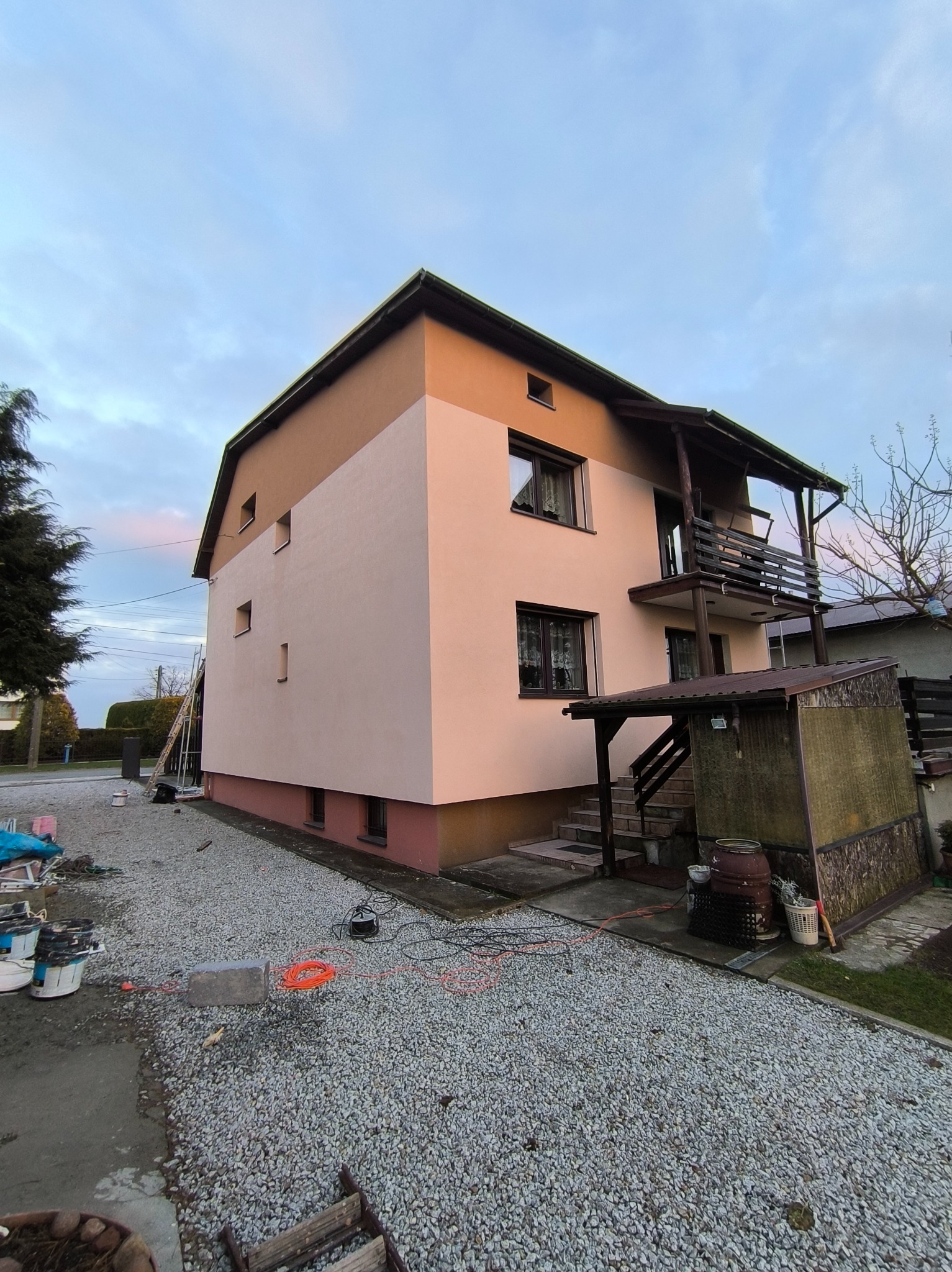 Dwukondygnacyjny dom po ociepleniu i malowaniu elewacji na beżowo-brązowe odcienie. Widoczne narzędzia malarskie i drabina. Otoczenie domu to żwirowe podłoże.