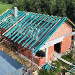 PAN OD DACHÓW SZYMON BIAŁECKI - Budowa domu jednorodzinnego z widoczną konstrukcją więźby dachowej pokrytą zieloną folią ochronną, ceglana elewacja w trakcie wznoszenia, widok z góry na plac budowy.