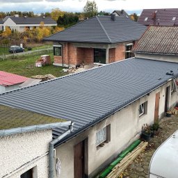 PAN OD DACHÓW SZYMON BIAŁECKI - Szary dach z blachy trapezowej na budynku gospodarczym podczas montażu, widoczna drabina i pracownik w ciemnej odzieży, w tle domy jednorodzinne i jesienne drzewa.