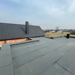 Widok z góry na fragment dachu pokrytego czarną papą bitumiczną na płasko, z kominem i fragmentem dachu skośnego z czarnej dachówki modułowej, w tle budynki mieszkalne.