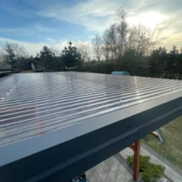 Przezroczysty dach z blachy trapezowej, mocowany śrubami, z widoczną konstrukcją wspornikową na tle ogrodu i błękitnego nieba.