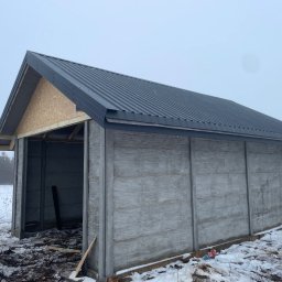 PAN OD DACHÓW SZYMON BIAŁECKI - Konstrukcja wiaty z prefabrykatów betonowych i blachy trapezowej na dachu, w zimowej scenerii, z widoczną niezabudowaną ścianą szczytową z płyty OSB.