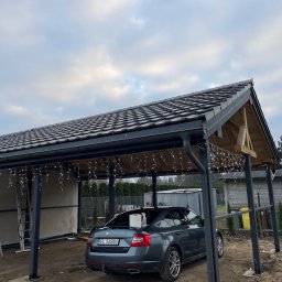 PAN OD DACHÓW SZYMON BIAŁECKI - Szara Skoda Octavia pod zadaszeniem z ciemną dachówką, ozdobionym lampkami, na tle pochmurnego nieba i ogrodzenia.