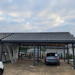 PAN OD DACHÓW SZYMON BIAŁECKI - Zadaszenie posesji z ciemną dachówką karpiówką, oświetlone girlandami, z samochodem na podjeździe i drabiny opartymi o ścianę budynku w tle pochmurne niebo.