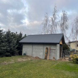 PAN OD DACHÓW SZYMON BIAŁECKI - Nowoczesny, szary budynek gospodarczy z blachy trapezowej na dachu, widoczny w ogrodzie z drzewami iglastymi i brzozami na tle pochmurnego nieba.