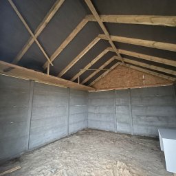 PAN OD DACHÓW SZYMON BIAŁECKI - Wnętrze nowo wzniesionej konstrukcji z drewnianą więźbą dachową pokrytą czarną membraną, szare betonowe ściany z widocznymi łączeniami, piaszczysta podłoga i biała szafka w rogu.