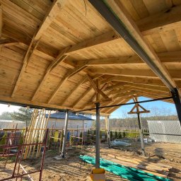 PAN OD DACHÓW SZYMON BIAŁECKI - Wnętrze nowo budowanego drewnianego zadaszenia z widoczną konstrukcją więźby dachowej i metalowymi słupami nośnymi, w tle zabudowa mieszkalna i drzewa.