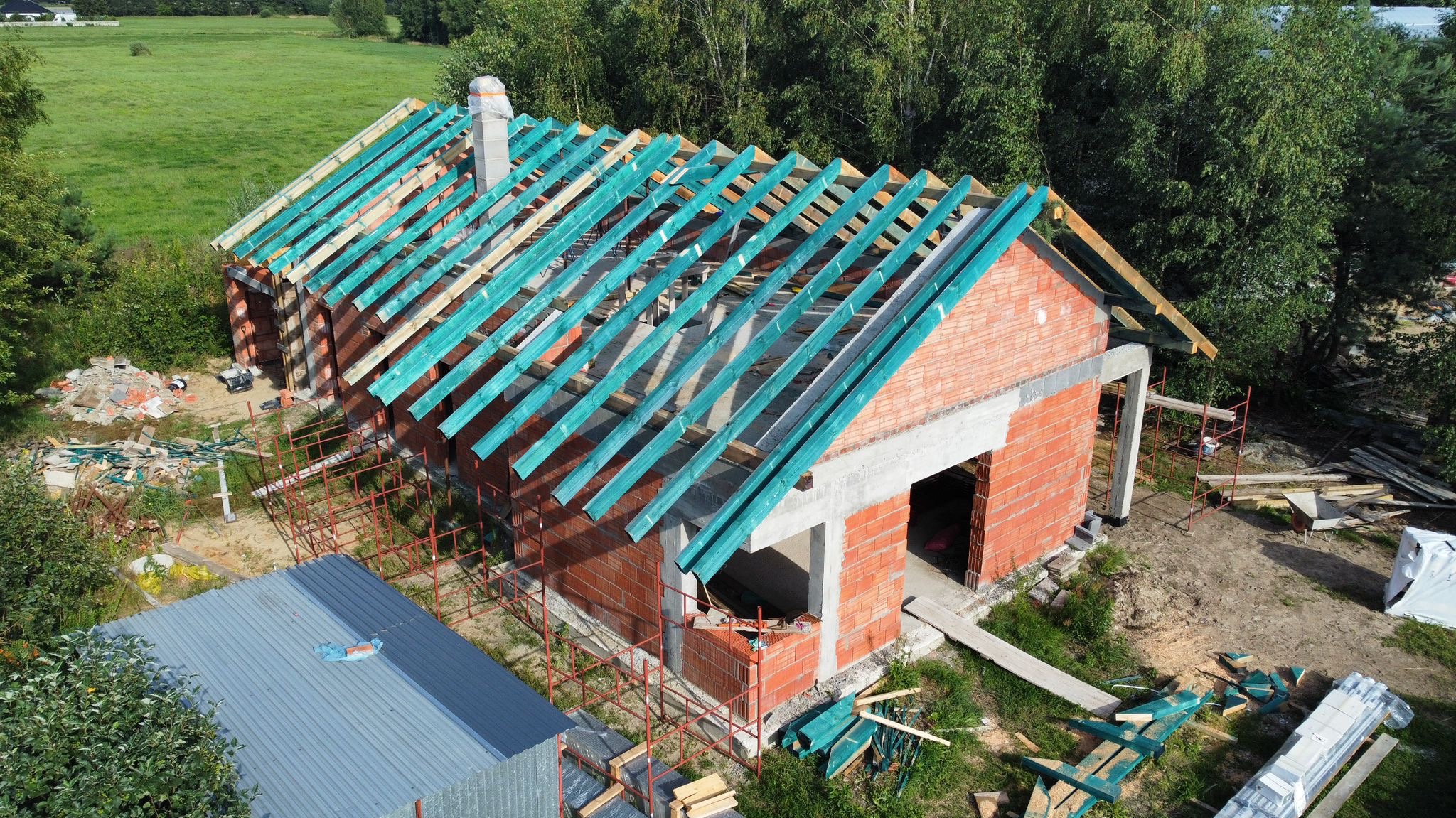 Budowa domu jednorodzinnego z widoczną konstrukcją więźby dachowej pokrytą zieloną folią ochronną, ceglana elewacja w trakcie wznoszenia, widok z góry na plac budowy.
