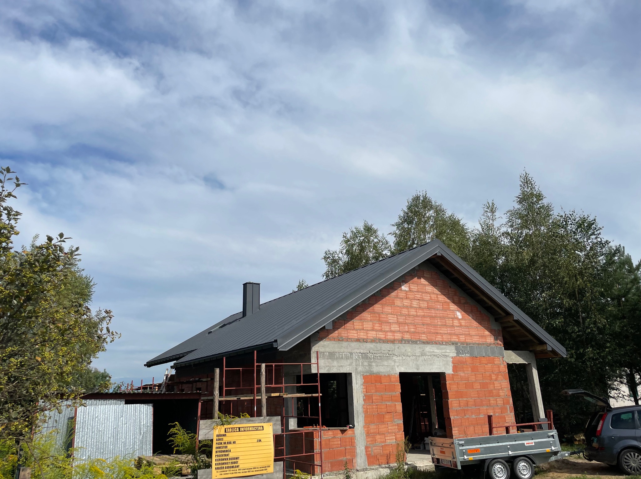 Dom w trakcie budowy z czarnym dachem i czerwoną cegłą, widoczna tablica informacyjna budowy i przyczepka samochodowa