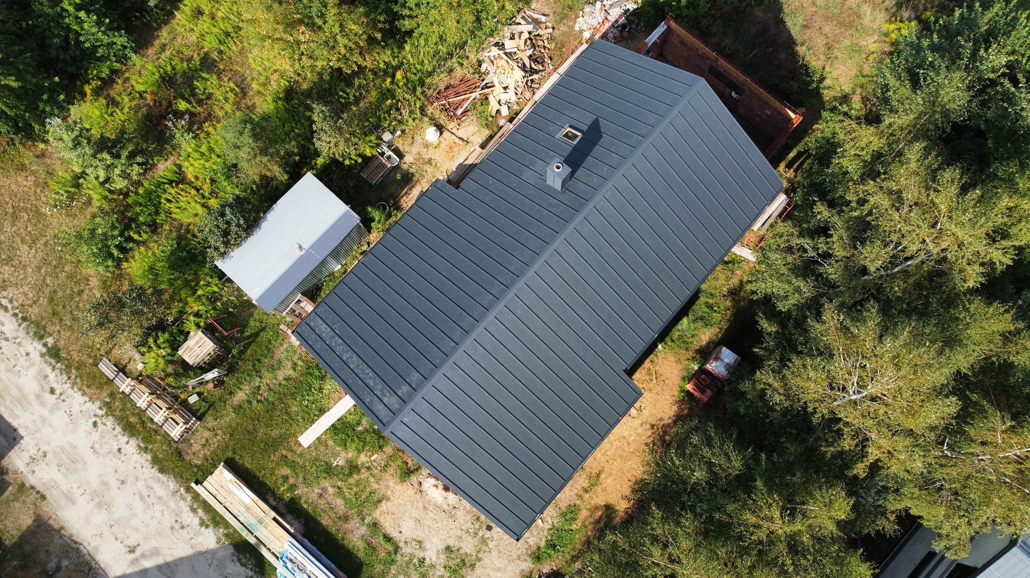 Widok z góry na dom z ciemnoszarym, metalowym pokryciem dachowym, kominem i oknem dachowym, otoczony zielenią i materiałami budowlanymi.