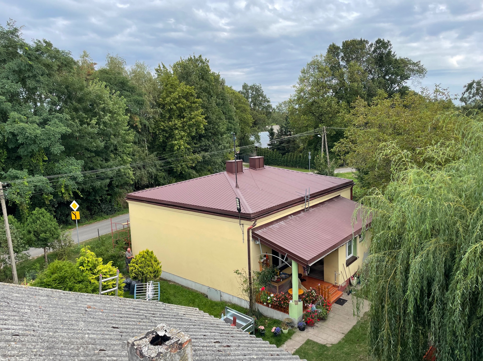 Dom jednorodzinny z nowym, bordowym dachem widziany z góry, z widoczną starą dachówką na pierwszym planie i pracującym robotnikiem przy elewacji w tle.