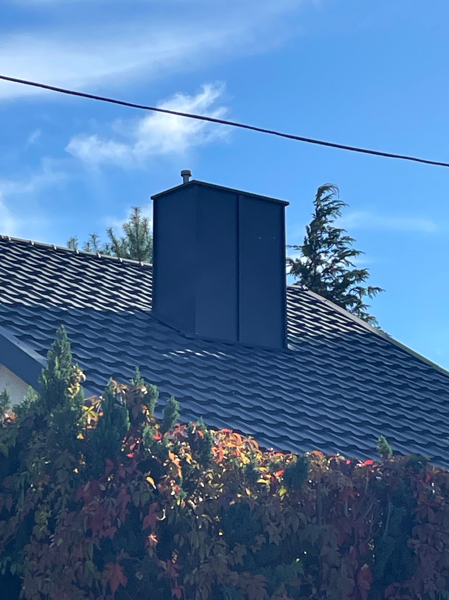 Nowoczesny, ciemny komin wentylacyjny na dachu pokrytym ciemną blachodachówką, widoczny na tle błękitnego nieba i zieleni drzew.