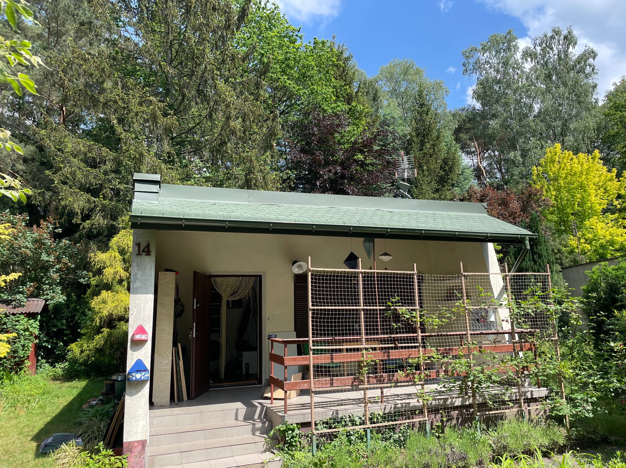 Niski dom z zielonym dachem krytym dachówką bitumiczną, numer 14 na filarze, antena telewizyjna na dachu, bujna roślinność w ogrodzie.